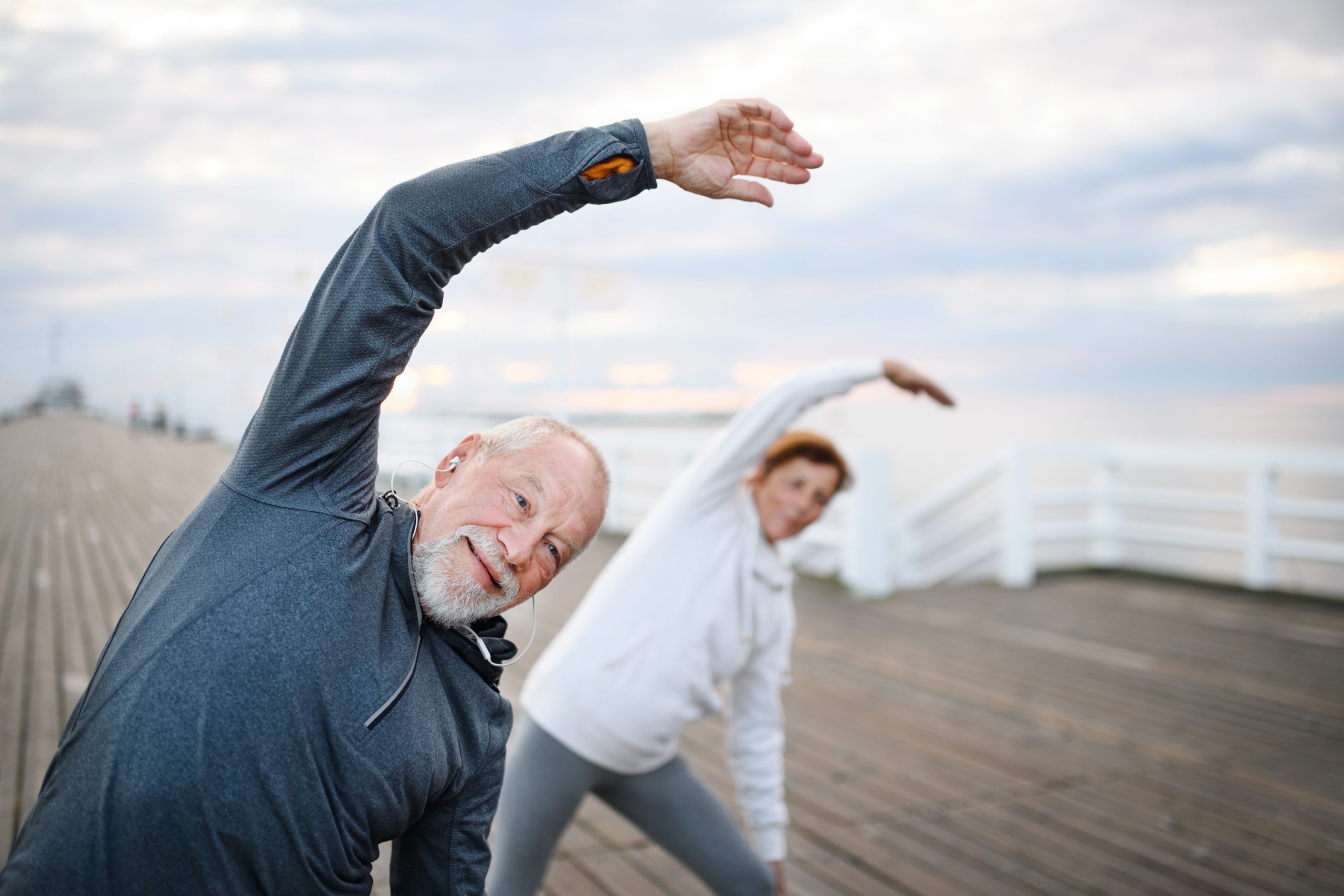 Active senior couple doing stretching exercise outdoors on pier by sea in early morning.