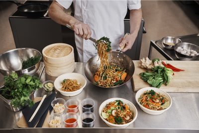 Kokki seoittamassa ruokaa pannulla ja annostelemassa sitä pöydällä oleviin take away kulhoihin