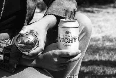 Mustavalkoisessa rajatussa kuvassa mies on kyykyssä petanque-pallon kanssa ja toisessa kädessä on Hartwall Vichy tölkki.