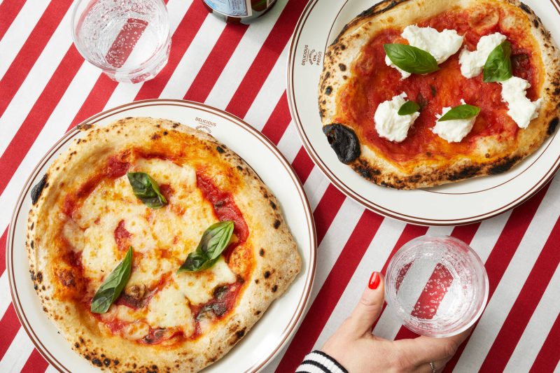 Kuvassa puna-valkoraidallisella pöytäliinalla käsi, kaksi vesilasia ja ja kaksi napolilaista pizzaa lautasilla.